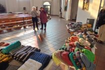 Einige Menschen stehen in einer Kirche und unterhalten sich, im Vordergrund sind drei Tische mit selbst gestrickten Dingen wie z.B. Schals und Mützen.