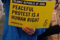 Person in blauer Regenjacke hält ein Amnestyplakat: "Islamic Republic of Iran: Peaceful protest is a human right".