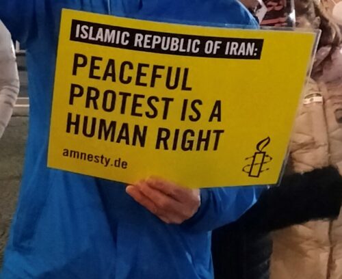 Person in blauer Regenjacke hält ein Amnestyplakat: "Islamic Republic of Iran: Peaceful protest is a human right".
