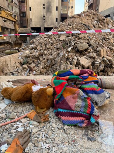 Eine bunte Wolldecke (von der Aachener Strickgruppe) liegt neben einem Teddybär vor den Trümmern eines Hauses in Beirut.