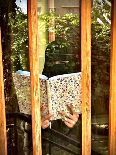 Eine Frau in einer Burka steht hinter einen Fensterscheibe in der sich die Aussenwelt speigelt und ließt in einem Buch.
