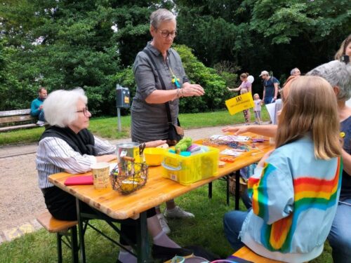 An einer Bierzeltgarnitur im Aachener Stadtpark zeigen Teilnehmer*innen des Strickcafés verschiedene Techniken. OB Sybille Keupen schaut vorbei.