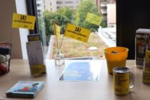Amnesty Infotisch mit Amnesty Journal, Flyern, AEMR für Kinder, Spendendose und Fähnchen mit Aufschrift "Ja! Zum Flüchtlingsschutz". Durch ein Fenster im Hintergrund sieht man Hochhäuser und ein begrüntes Dach.