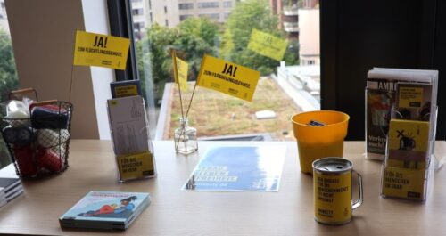 Amnesty Infotisch mit Amnesty Journal, Flyern, AEMR für Kinder, Spendendose und Fähnchen mit Aufschrift "Ja! Zum Flüchtlingsschutz". Durch ein Fenster im Hintergrund sieht man Hochhäuser und ein begrüntes Dach.