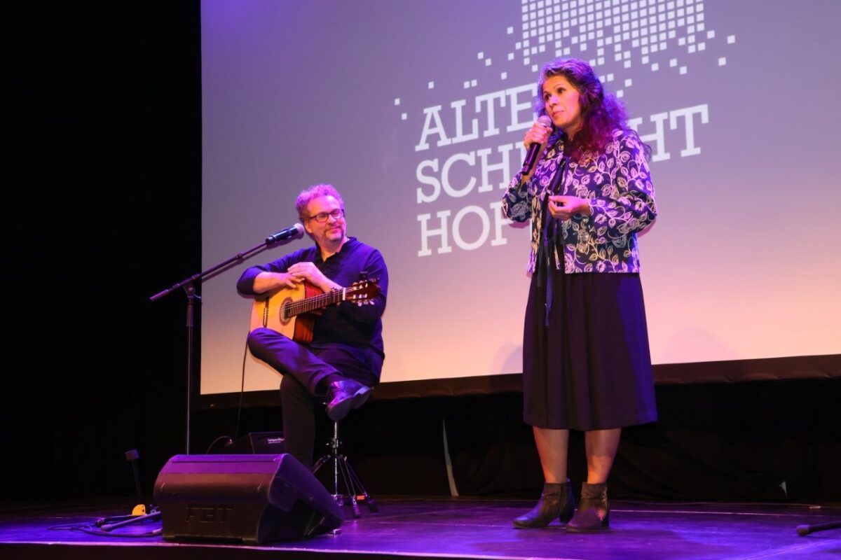 Auf der Bühne des "Alten Schlachthof" steht eine Frau mit einem Mikrofon, daneben sitzt ein Mann mit einer Gitarre.
