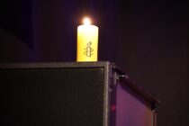 Eine gelbe Amnesty-Kerze brennt vor dunklen Hintergrund. Sie steht auf einer Lautsprecherbox.