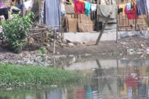 Im Vordergrund ein Gewässer, dahinter schließt sich ein Gelände mit einfach gemauerten Hütten und Wellblechdächern an. Auf Wäscheleinen hängen verschiedene, bunte Wäschestücke zum trocknen.