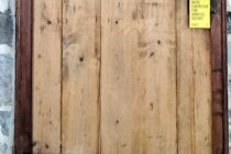 Tür aus alten Holzbrettern mit schwarz lackierten Holzbeschlägen. An der Klinke hängt ein gelbes Schild mit Aufschrift "Bitte eintreten für Arbeitsrechte".